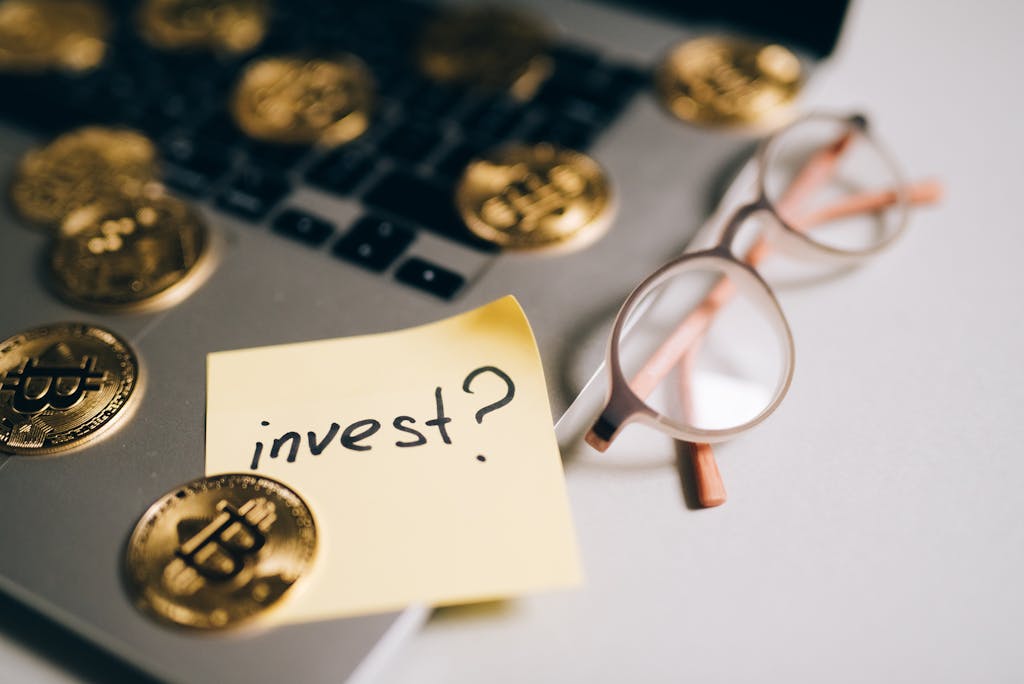 A close-up of bitcoin coins on a laptop with eyeglasses and a sticky note reading 'invest?'